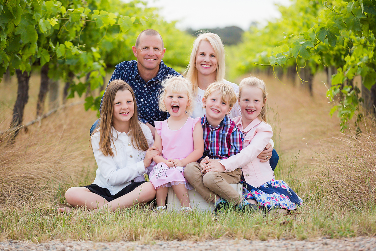 mum and dad and their four children laughing and smiling on a rug in the vineyards at their family photo session by deb elton photography